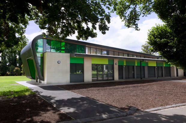 A contemporary single-storey institutional or commercial building set within landscaped grounds. The long facade combines pale concrete or rendered wall panels, extensive dark-framed glazing, bright green and yellow-green feature panels, and a distinctive curved or barrel-like glazed end volume. A paved pedestrian path leads toward the covered entrance frontage, with mature trees surrounding the site.