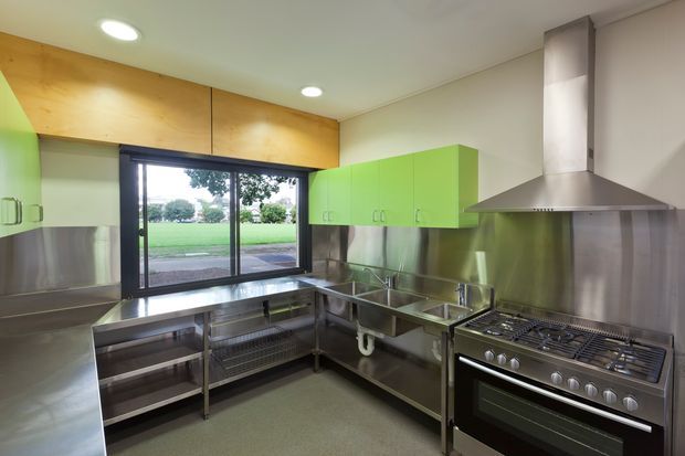 A commercial-style stainless-steel kitchen with extensive open shelving, stainless work surfaces, double sinks, a large gas range and industrial canopy hood. Bright lime-green overhead cabinetry contrasts with brushed metal surfaces, while a timber-lined wall and large glazed door provide warmth and views to a lawn outside.