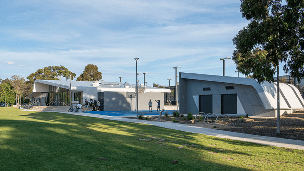A contemporary low-rise civic or educational complex set within a landscaped park. The foreground is a broad lawn with mature trees, while the buildings feature angular forms, pale grey cladding or concrete-like facades, dark recessed openings, extensive glazing, and simple modern light poles. A paved pedestrian path and bicycle racks run along the building frontage. The image is an exterior architectural view in clear daylight.