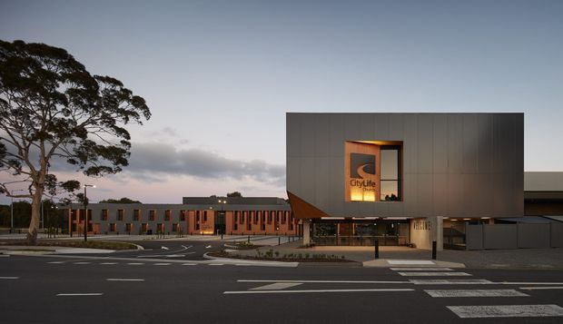 Exterior architectural view of a contemporary civic or commercial building at dusk. A large dark rectangular upper volume projects over a glazed entrance, with a vertically illuminated sign and warm interior lighting. The surrounding low-rise brick and dark-clad buildings, broad road markings, pedestrian crossings, streetlights, and mature tree establish an urban institutional setting.