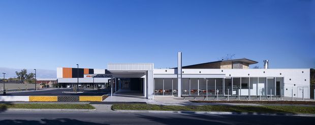 Wide exterior view of a low-rise contemporary commercial building with a mostly white facade, extensive glazed frontage, a projecting flat roof, and a covered recessed entry or loading-style opening. The building sits along a paved street with minimal landscaping and a clear blue sky.