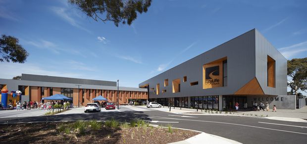 Wide exterior view of a large contemporary community or commercial building with a sharply angled roofline, grey metal-clad facade, warm timber-toned feature panels, extensive ground-floor glazing, and a prominent welcome entrance. A forecourt and broad paved roadway occupy the foreground, with people and vehicles near the building.