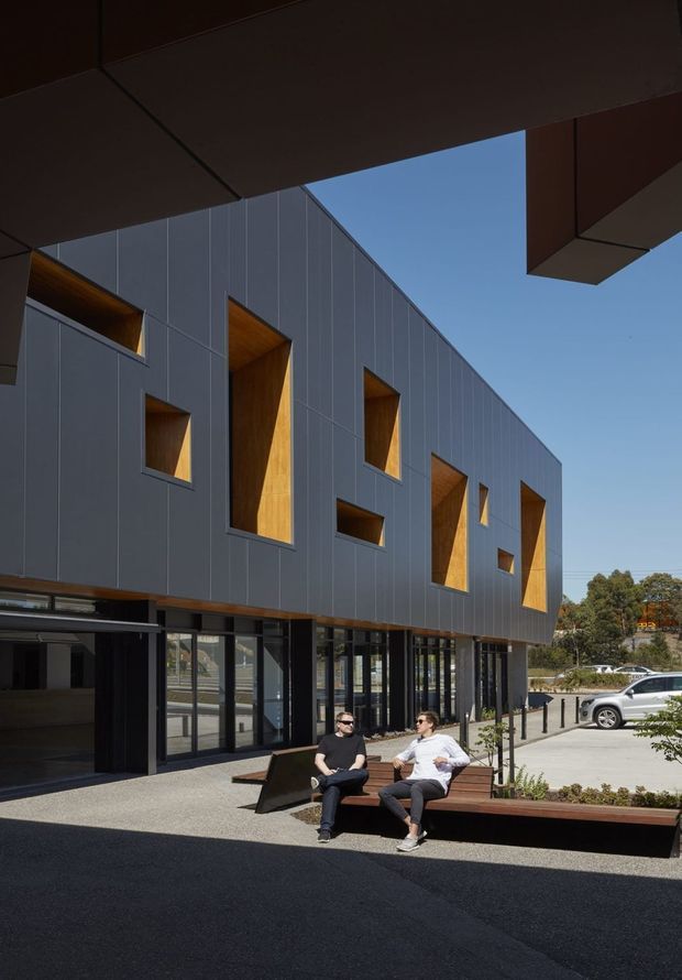 Exterior architectural photograph of a contemporary multi-storey building with a dark grey panel-clad upper façade punctuated by deep, irregularly arranged rectangular openings lined in warm timber. The glazed ground floor appears commercial or institutional, opening onto a paved public forecourt with integrated timber bench seating and people. Large dark overhead canopy elements frame the view, with a clear blue sky and parking area beyond.