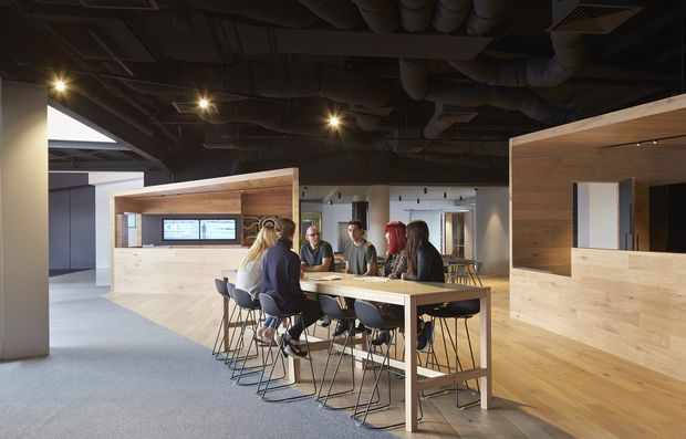 Open-plan commercial office interior with a communal high table and stools occupied by several people, surrounded by large light-wood veneered partition and reception-like joinery. The space has exposed dark ceilings with visible ductwork and services, grey carpet, integrated linear lighting, and a contemporary collaborative workplace layout.