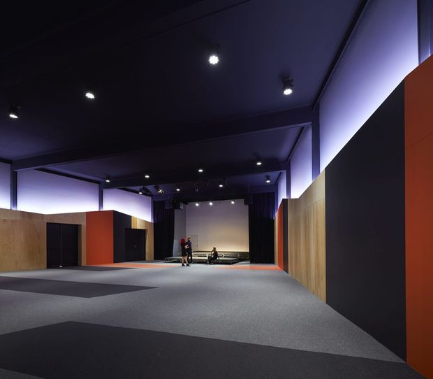Large, dark contemporary interior resembling a gallery, theatre foyer, or event space, with a broad carpeted floor, black ceiling, recessed and track lighting, and sculptural partition walls finished in timber, black, and orange-red panels. A small stage or presentation area with curtains and seating is visible at the far end. The space has dramatic purple accent lighting and a highly open, spacious layout.