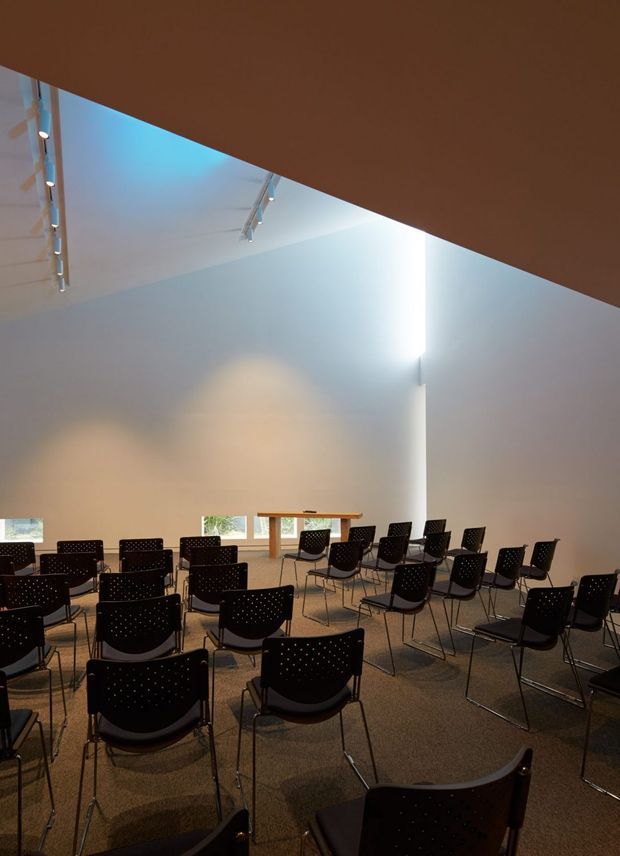 A spacious, high-ceilinged multipurpose room arranged with rows of black perforated chairs facing a simple timber lectern or presentation table. The room has plain light walls, grey carpet, narrow horizontal windows, exposed track lighting, and dramatic indirect illumination along the ceiling and wall junctions. The composition suggests a lecture, meeting, assembly, or event space.
