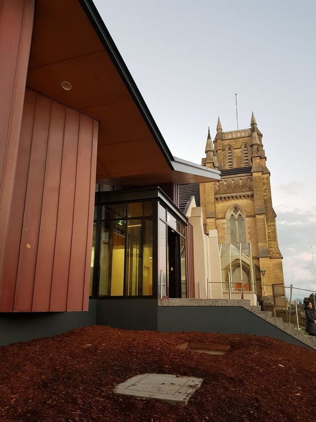Exterior architectural photograph showing a contemporary building extension with reddish-brown vertical cladding, deep projecting roof/eave, dark base, and extensive glazed entry or lobby frontage. A historic stone church with Gothic Revival features, arched windows, pinnacles, and tower rises immediately behind the modern addition. The foreground contains landscaped mulch and a paved access area.