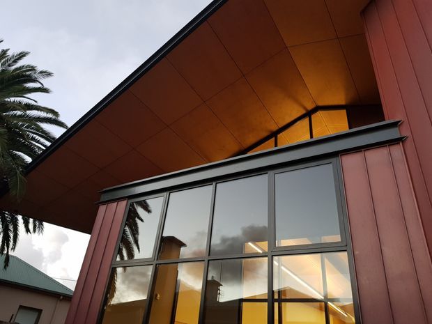 Upward-looking exterior view of a contemporary building with a dramatic deep roof overhang and warm timber-lined soffit. Large dark-framed floor-to-ceiling glazing reflects palm trees and the cloudy sky, while vertical reddish metal-clad wall panels frame the facade. Warm interior lighting is visible through the windows.