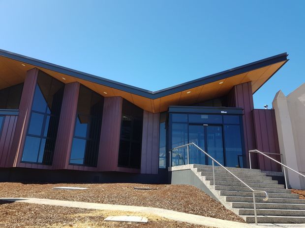 Exterior view of a contemporary public or commercial building with a dramatic V-shaped roofline, deep timber-toned soffits, tall dark glazing, and vertical burgundy cladding panels. A broad stair and accessible entry ramp lead to a glazed entrance, with landscaped gravel and concrete paths in the foreground under clear daylight.