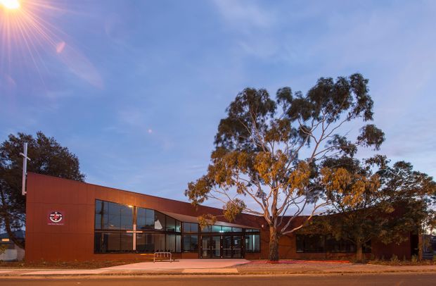 Low, contemporary commercial building viewed from across the street at sunset. The facade combines warm reddish-brown metal cladding with extensive dark-framed glazing, a recessed glazed entrance, and a shallow flat roofline. Mature eucalyptus trees partially screen the building, while warm interior lighting creates a welcoming evening appearance.