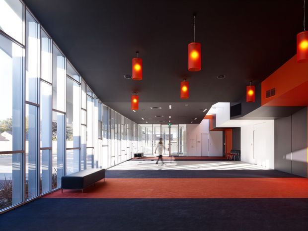 A large, modern institutional interior hall with floor-to-ceiling glazed curtain walls along one side, a dark ceiling, vivid red pendant lights, red and charcoal carpet, and white built-in wall elements. Strong linear daylight and geometric shadow patterns extend across the spacious circulation or gathering area.