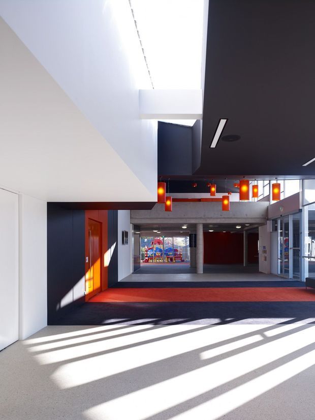 Bright contemporary interior circulation space, likely a school or institutional foyer, with dramatic clerestory daylight, white and charcoal architectural surfaces, concrete structural elements, red carpet zones, vivid orange pendant lights, and extensive glazing to the right. The deep view leads toward a colourful children’s play area or artwork beyond.