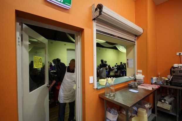 A compact commercial food-preparation or service area with vivid orange painted walls, a stainless-steel workbench, commercial coffee equipment, shelving, and a large internal service opening with a roller shutter. A glass door and service window reveal a busy adjoining room with people and tables. The space is functional and brightly lit, with exposed equipment and storage supplies.