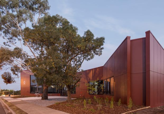 Low-rise contemporary building with a bold dark red-brown metal-clad facade, prominent pitched roof form, and large glazed openings. The building is set within a landscaped site with mature eucalyptus trees, gravel, mulched planting beds, and a concrete pedestrian path. Warm late-day light highlights the facade and surrounding vegetation.