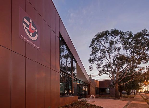 Exterior architectural photograph of the Arthur W. White University Centre, showing a contemporary institutional building clad in reddish-brown rectangular panels. Tall glazed openings reflect surrounding eucalyptus trees, while a paved pedestrian forecourt and mature tree frame the low, angular building at dusk.