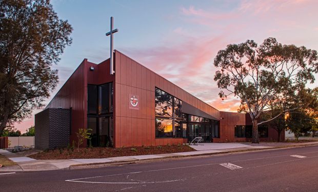 Exterior view of a contemporary single-storey red-brown metal-clad building with a steep asymmetrical gabled roof and prominent cross, likely a church or community facility. Large glazed openings reveal warm interior lighting, while mature trees and a paved street frame the dusk scene.