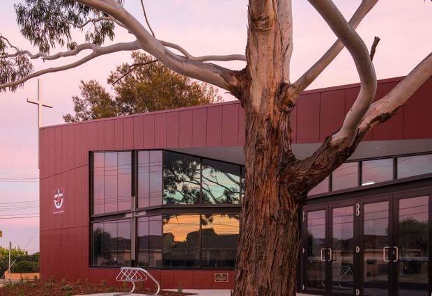 Exterior view of a contemporary low-rise building clad in deep burgundy-red panels, with extensive dark-framed glazing and a glazed entrance. A large mature eucalyptus tree dominates the foreground, while a simple cross and circular emblem suggest the building may be a community or religious facility. The image is photographed at dusk with warm reflections in the windows.