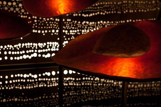 A dark, atmospheric interior installation featuring layered, organically shaped copper or rust-toned metal canopy forms with warm orange illumination. Perforated dark wall and ceiling surfaces create dense patterns of glowing circular openings, producing dramatic reflections and a sculptural, immersive ambience.