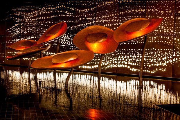 A dramatic art installation in a dark interior, featuring several large sculptural forms resembling glowing orange-red flowers or abstract discs on slender stems. The installation stands on a highly reflective dark tiled floor, with a perforated or dotted metallic backdrop creating shimmering patterns of light and reflections. The atmosphere is immersive, theatrical, and warm-toned.