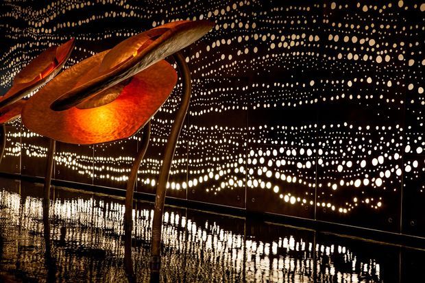 A dark immersive interior installation with mirrored black walls and ceiling covered in dense patterns of circular light points, creating a star-like reflected environment. A large sculptural orange-red metallic form with curved supports dominates the foreground, standing on a highly reflective dark floor. The scene is dramatic, artistic, and gallery-like.