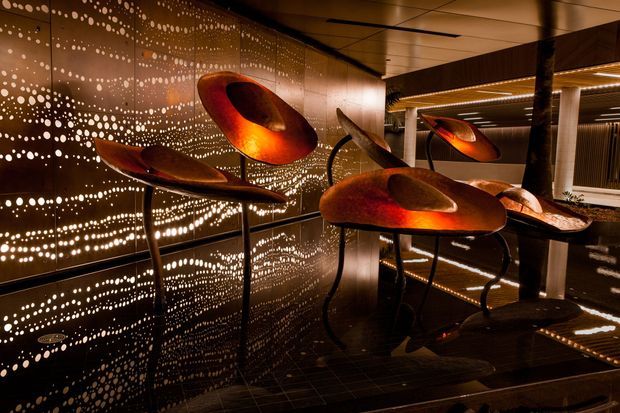 A dark, highly stylised interior exhibition or commercial display space featuring sculptural copper- and bronze-toned abstract objects on slender black stands. Perforated metallic wall panels create dense dotted reflections, while linear recessed lighting and glossy dark surfaces produce a dramatic, immersive atmosphere.