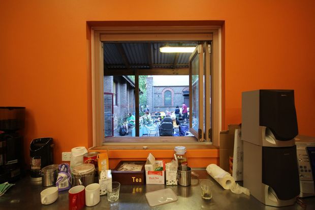 A compact cafe or kitchenette workspace viewed from inside, with vivid orange walls, a timber-framed open serving window, stainless-steel counter, coffee machine, cups, tea supplies, jars, and other beverage-making equipment. Through the window, an exterior courtyard or laneway with brick buildings and bicycles is visible. The scene has a utilitarian, eclectic commercial interior feel.