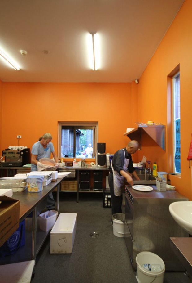 A compact commercial kitchen or food-preparation workspace with vivid orange painted walls, stainless-steel work benches and sinks, open shelving, cooking equipment, fluorescent ceiling lights, and two people preparing food. The room has a practical, utilitarian fit-out with dark flooring and several windows.
