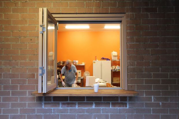 A small service-style window opening through a dark red-brown brick wall, with timber-framed casement shutters opened outward and a deep timber sill. Through the opening is a compact orange-painted interior resembling a workshop, utility room, or kitchenette, with shelving, a refrigerator, countertop items, and a person. The image is documentary and functional, with warm orange and brick tones.