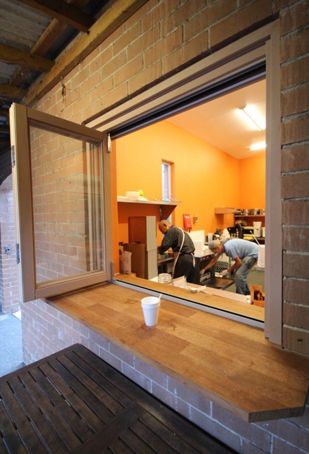 View through an open service window into a small commercial kitchen or food-preparation area. The foreground has a timber-framed opening with a broad timber counter and dark timber decking below. Exposed orange-toned brick walls and exposed timber roof framing surround the opening, while the brightly painted orange interior contains workers, stainless-steel equipment, shelving, and food-service items.