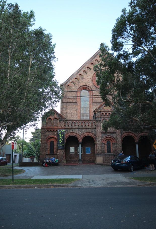 Street-level exterior view of a historic red-brown brick church or institutional building, partly screened by mature trees. The facade features a steep gable, repeated round-arched brick detailing, a large central arched window, and a recessed arcaded entrance supported by columns. A paved forecourt, street frontage, signage, pedestrians, and parked cars are visible.