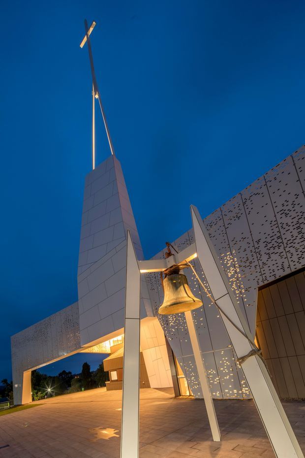 Dramatic dusk exterior view of a contemporary civic or commercial building, featuring a tall tapered tiled spire with a cross-like finial, perforated metal facade panels, angular white structural blades, and a prominent suspended bronze bell. The paved forecourt and illuminated building create a striking architectural landmark composition against a deep blue sky.