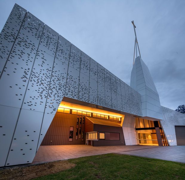 Exterior dusk view of a contemporary institutional or commercial building with a dramatic angular facade. Large pale metal panels are perforated with irregular horizontal slots, while warm illuminated recessed entrances and timber-toned cladding contrast with the cool exterior. A sharply pointed tower-like architectural element rises above the entrance, and paved forecourt and lawn occupy the foreground.