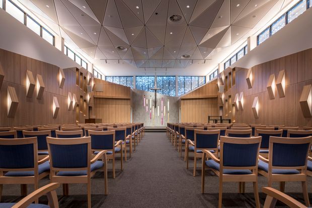 A spacious contemporary auditorium or chapel-like gathering hall viewed down the central aisle, with rows of blue upholstered timber-framed chairs, warm wood wall panelling, geometric wall sconces, a textured angular ceiling with recessed lighting, and high clerestory glazing. A feature wall and raised focal area are visible at the far end.