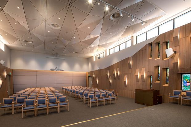 A spacious contemporary chapel or ceremonial hall with rows of blue upholstered chairs, a raised front platform, warm timber wall panelling, narrow stained-glass windows, geometric wall lighting, and an angular acoustic ceiling with recessed spotlights. The room is bright, orderly, and designed for communal gatherings or services.