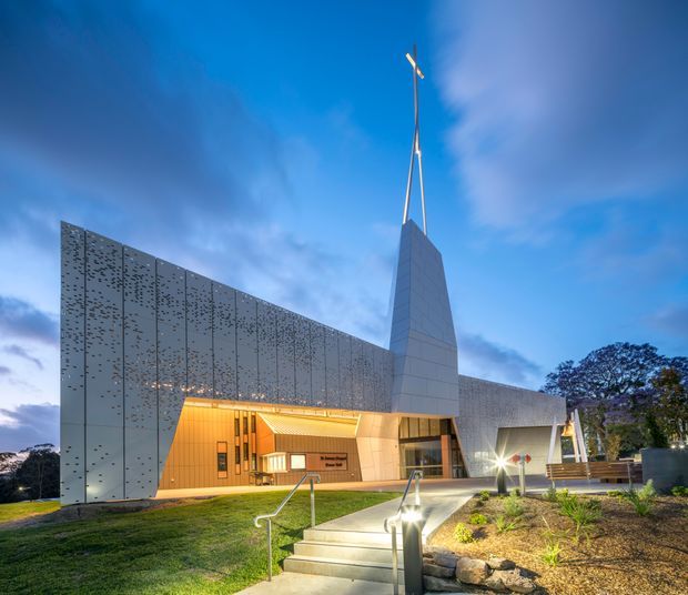 Exterior architectural photograph of a contemporary institutional building at dusk. The building features a large perforated metal-clad facade, a projecting angular entry canopy with warm interior lighting, a tall tapered tower or spire, and landscaped pedestrian access with steps and handrails.