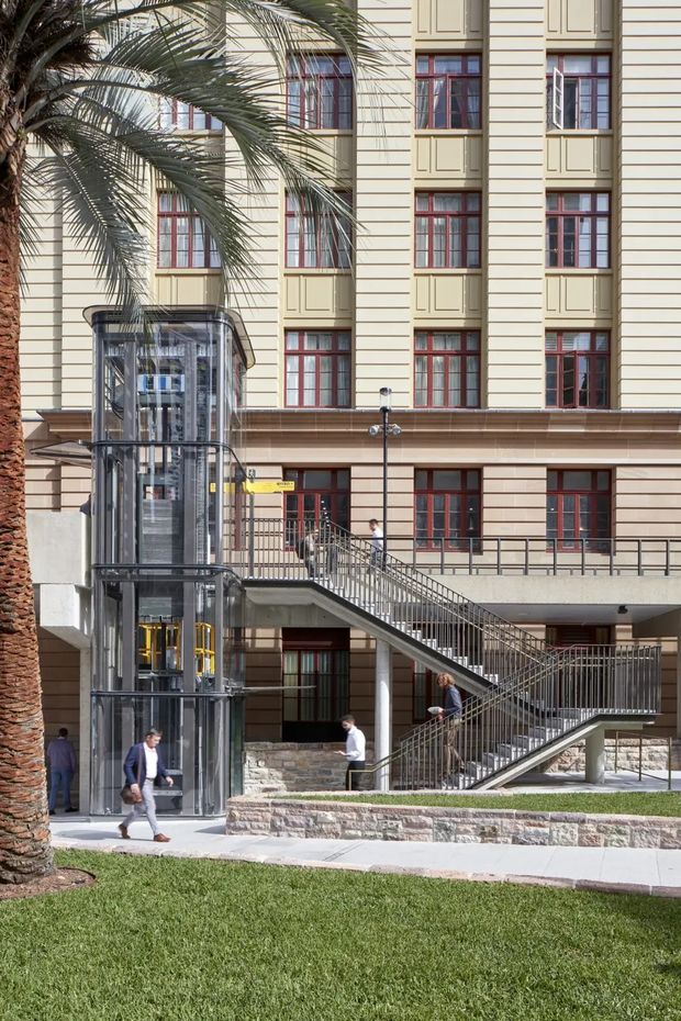 Exterior view of a multi-storey institutional or commercial building with a cream horizontal-panel facade, recessed window bays, and dark red timber-framed windows. A glazed external elevator shaft with visible metal structure stands beside an accessible-looking stairway and ramp circulation system. The foreground includes a landscaped lawn, palm tree, paved paths, stone retaining walls, and several pedestrians.
