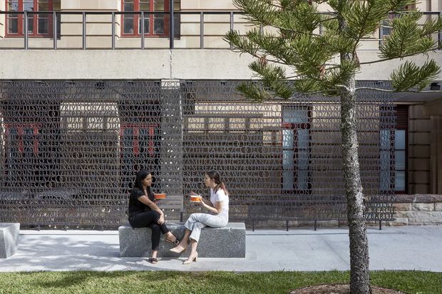 Exterior view of a contemporary multi-storey building with a distinctive perforated metal screen wall covered in layered lettering. Two people sit on a simple stone or concrete bench along a paved forecourt, with trees and grass in the foreground. The upper facade has rectilinear window bays with dark frames and muted red accents.