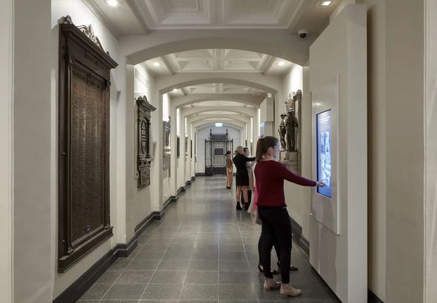 A long, formal institutional corridor with repeated arched openings, coffered and beam-like ceiling detailing, pale plaster walls, dark tiled flooring, and dark decorative wall panels and framed displays. Visitors are interacting with a digital information screen along the right wall, while statues and exhibits line the passage. The space reads as a museum or heritage gallery interior.