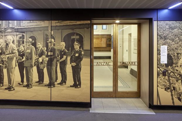 Entrance to a commemorative museum or exhibition space, with timber-framed glass double doors labelled “ANZAC SQUARE,” surrounded by large sepia historical photographic wall panels. The interior has dark blue-black walls, a tiled threshold, and a ribbed ceiling with linear lighting.