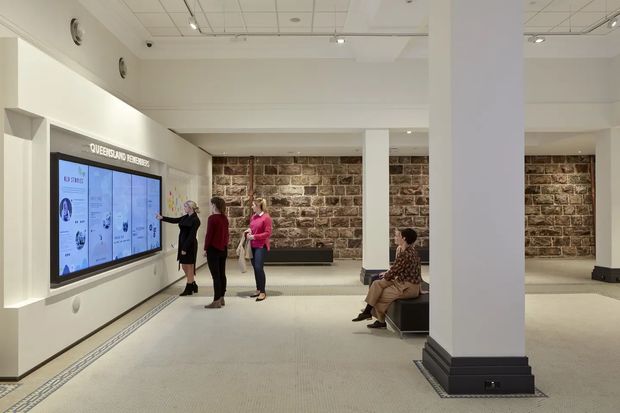 Bright, spacious institutional or commercial interior with a large interactive digital display, visitors viewing information, built-in bench seating, substantial square columns, and exposed rough stone feature walls. The room has a suspended acoustic ceiling, light carpeted flooring, and a clean gallery or reception-like layout.
