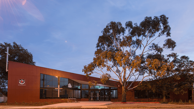 Single-storey contemporary institutional or commercial building with a low, rectilinear form and a rust-red metal-clad facade. Extensive dark-framed glazing forms the entrance and front elevation, with warm interior lighting visible through the windows. A mature eucalyptus tree partially screens the building, and a paved pedestrian approach runs along the front. The image is an exterior architectural photograph under a clear blue sky.
