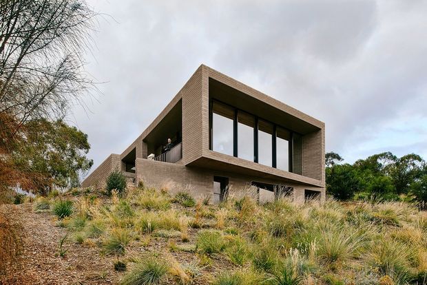 Low-angle exterior view of a contemporary, rectilinear house set into a landscaped sloping site. The building has a strongly projecting upper volume, a flat roof, extensive dark-framed floor-to-ceiling glazing, and textured brown masonry or brick walls. Native grasses and established planting surround the structure beneath an overcast sky.