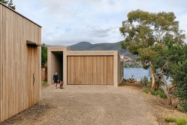 A contemporary timber-clad building complex viewed from a gravel driveway, with a large timber garage door and matching vertical wood siding. The site overlooks a body of water and wooded hills, with mature trees framing the scene and a person with a dog providing scale.