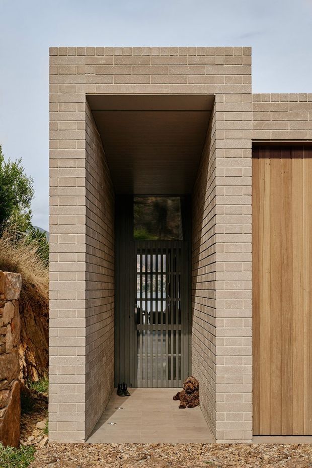 Exterior architectural view of a contemporary brick-clad entry passage. A deep, covered pedestrian breezeway is framed by pale beige brick walls and a timber-lined ceiling, leading to a dark vertical-slat gate. A tall timber door is visible at right, with rugged natural landscaping and a tree stump beside the structure.