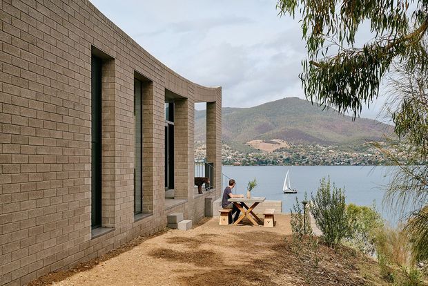 A modern curved masonry-clad building exterior beside a lake, with tall recessed windows overlooking the water and mountainous terrain. A small timber outdoor table and stools sit on a rough gravel terrace, partially screened by eucalyptus vegetation.