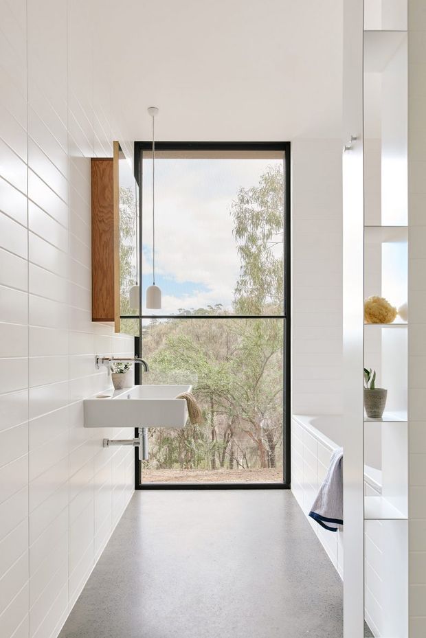Bright, narrow contemporary bathroom with white tiled walls, a wall-mounted rectangular basin and black tapware, a freestanding or built-in bathtub partially visible on the right, and open white shelving. A large black-framed window provides a view of trees and natural landscape. The space has a clean, minimal design with light grey polished concrete flooring, warm timber joinery, and simple pendant lighting.