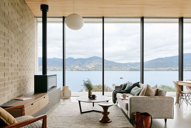 A bright contemporary living room overlooking a lake and mountainous shoreline through full-height black-framed glazing. The room features a pale exposed brick wall, warm timber-lined ceiling, light timber built-in media cabinetry, a sculptural black fireplace flue, neutral sectional sofa, round coffee tables, and minimalist pendant lighting.