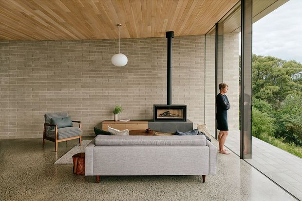A refined contemporary living room with a low-profile grey sofa and armchair arranged around a compact fireplace. The room features a warm timber-lined ceiling, exposed light-grey brick walls, a polished terrazzo floor, and expansive floor-to-ceiling glazing overlooking leafy greenery. A black freestanding wood-burning fireplace and simple pendant light provide focal points.
