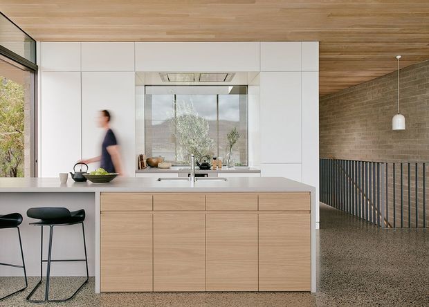A bright, minimalist open-plan kitchen with a large pale timber island, white flat-panel cabinetry, integrated appliances, and a central sink. The kitchen has a warm timber-lined ceiling, polished concrete floor, expansive glazing, black bar stools, and a black metal balustrade overlooking an adjacent lower level or stair void.