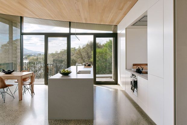 Bright contemporary open-plan kitchen and dining area with a central white island, extensive white built-in cabinetry, integrated appliances, and a timber-lined ceiling. Full-height dark-framed glazing opens to a balcony and leafy suburban views, while a polished speckled terrazzo floor continues through the space.